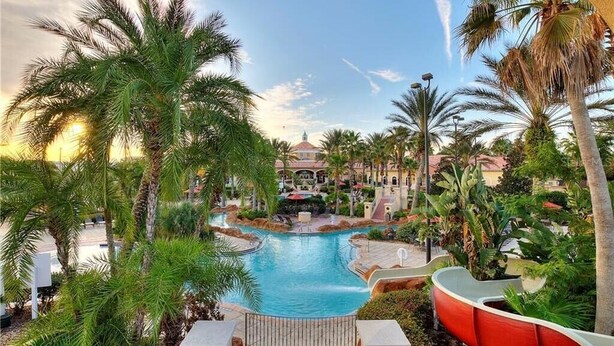 Zwembad van een resort met palmbomen, een waterglijbaan en een gebouw op de achtergrond.