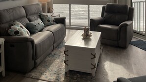 A modern living room with a large window offering a view of the ocean, a ceiling fan, a gray sofa with patterned cushions, a white coffee table, and a dark armchair. - Stunning Top-Floor 3 Bedroom, 2 Bath Oceanfront Condo 902 Pinnacle (North Myrtle Beach)