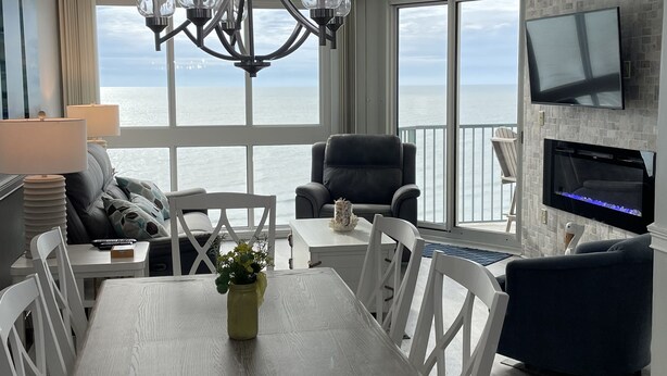 A modern living room with a dining area, large windows with a view of the ocean, a chandelier, and a flat-screen TV mounted on the wall. - Stunning Top-Floor 3 Bedroom, 2 Bath Oceanfront Condo 902 Pinnacle (North Myrtle Beach)