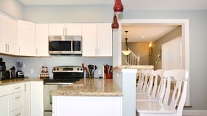 A kitchen with white cabinets, a stainless steel microwave, a stove, and a granite countertop. There is a dining area with white chairs and a table. - Pet Friendly Downtown Rehoboth Beach House (Rehoboth Beach)