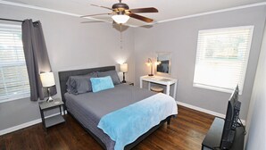 Un dormitorio con cama, ventilador de techo, mesita de noche con lámpara, cómoda, espejo y televisor sobre un mueble.