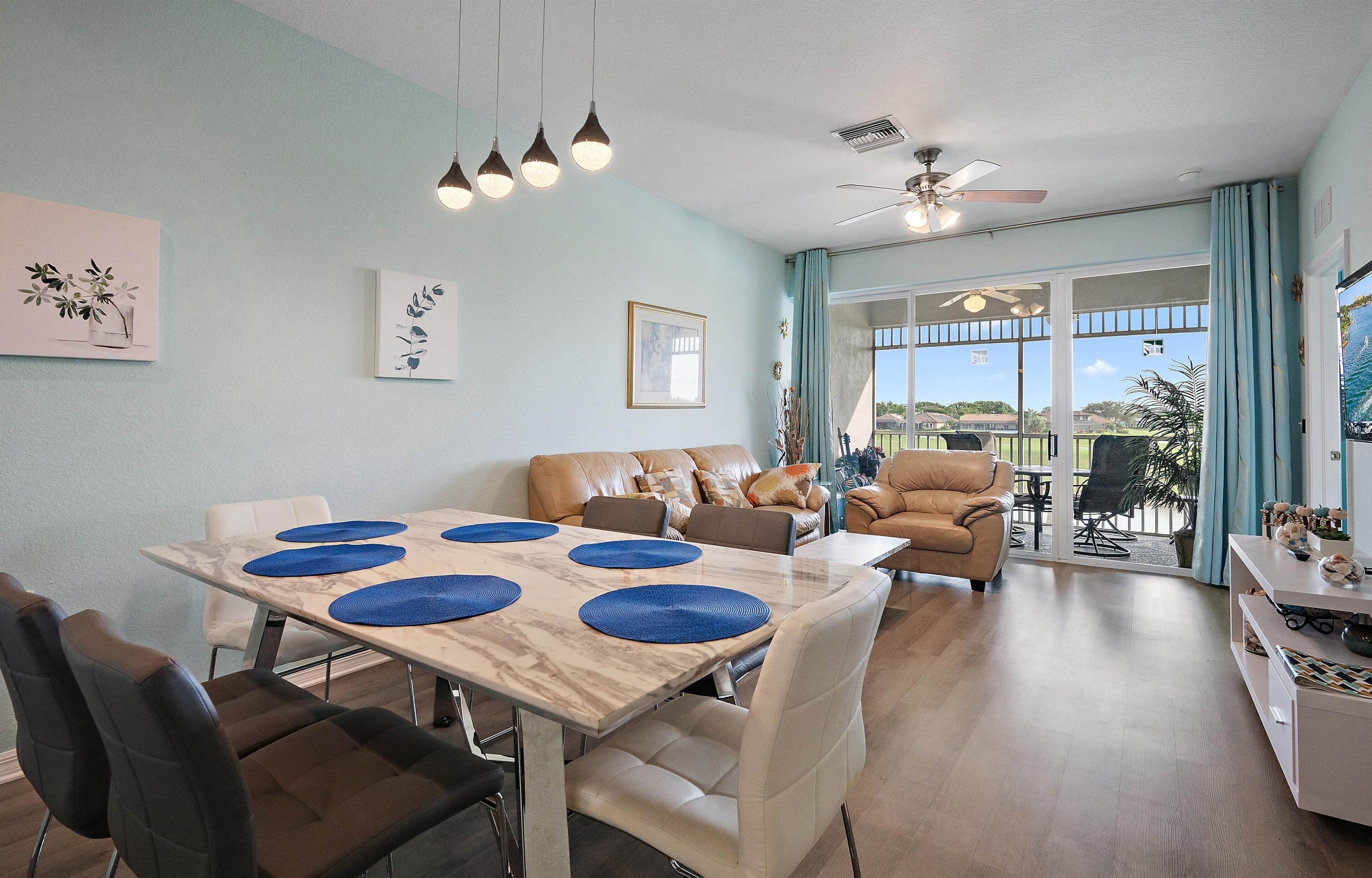 A spacious living room with a dining area, a ceiling fan, and large windows offering a view of the outdoors.