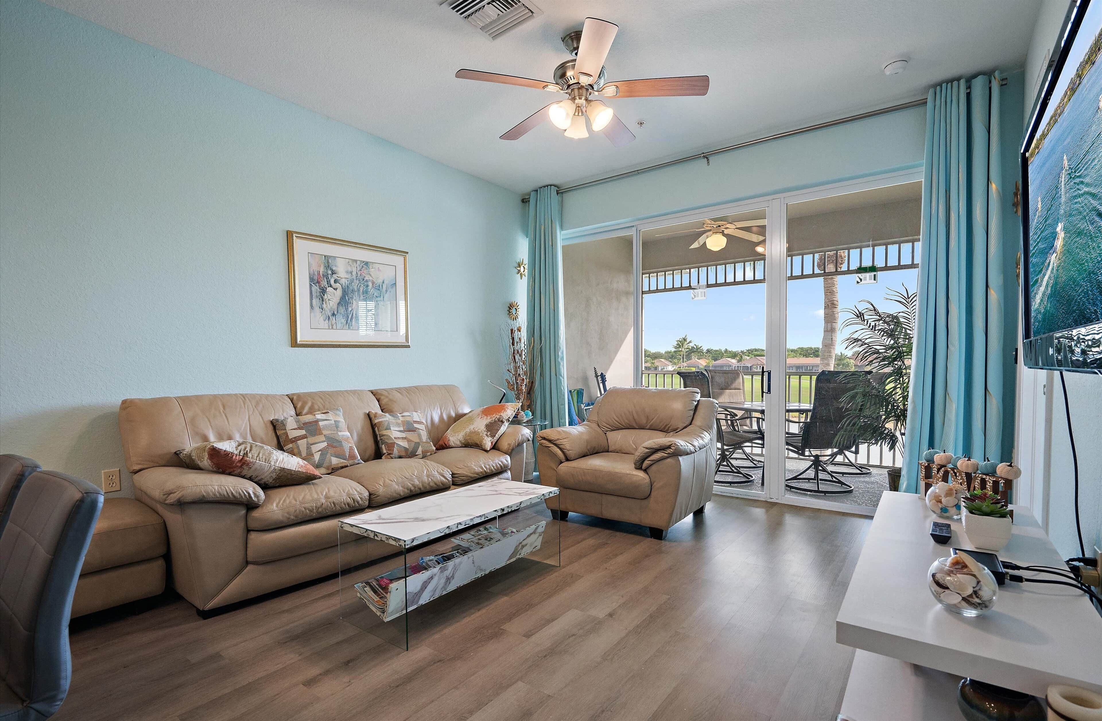 A living room with a beige sofa, a coffee table, a chair, and a TV on a stand. There is a ceiling fan, a window with curtains, and a view of the outdoors.