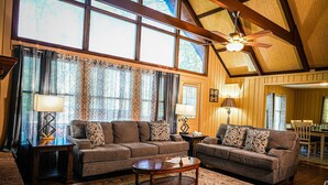 A spacious living room with two sofas, a coffee table, and a ceiling fan. Large windows allow natural light to fill the room.