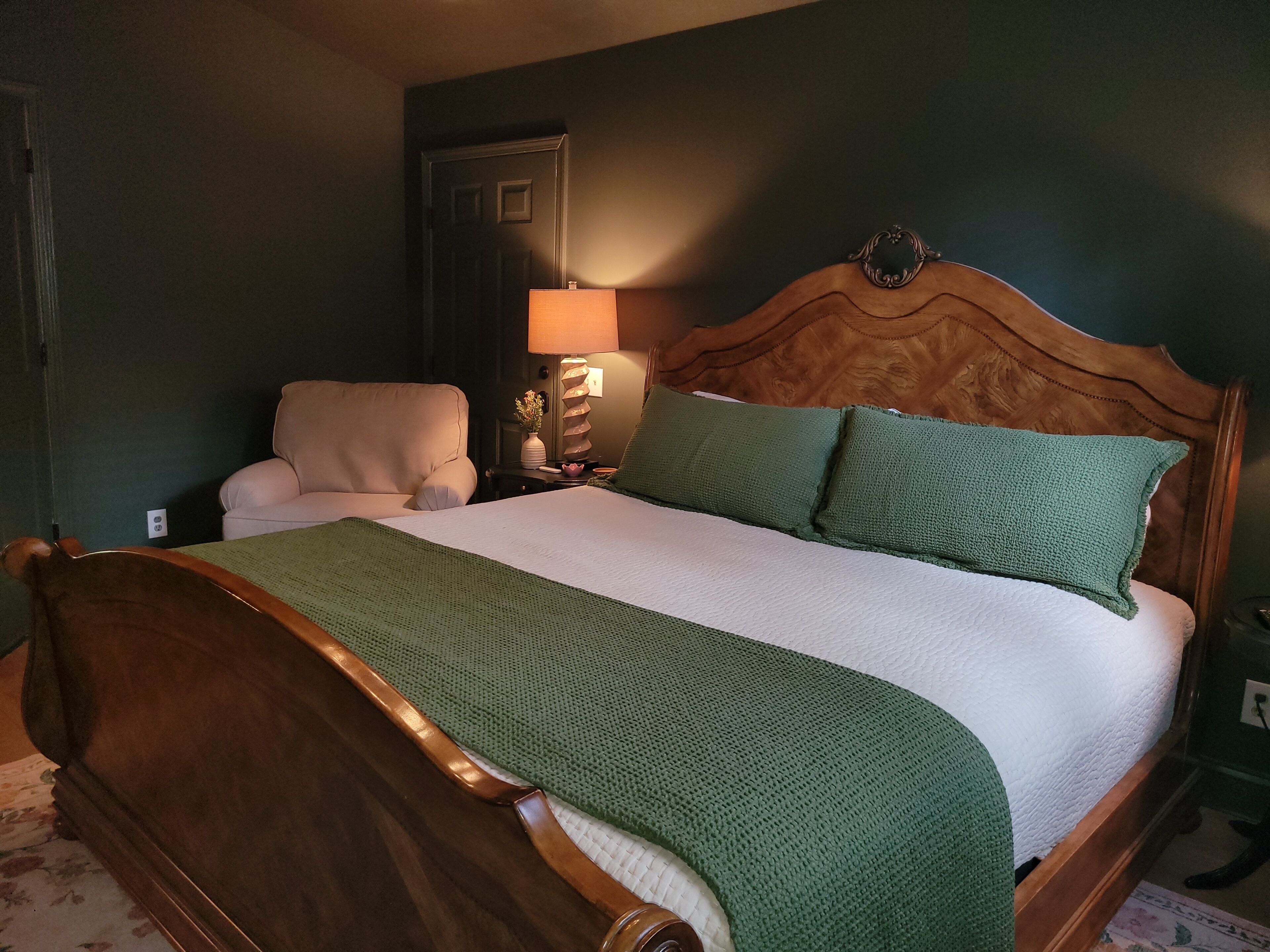 A bedroom with a wooden bed, a green throw blanket, a bedside lamp, and a chair.
