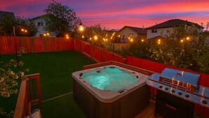 Un jardin avec un jacuzzi, un barbecue et des guirlandes lumineuses, le tout illuminé par le ciel au coucher du soleil.