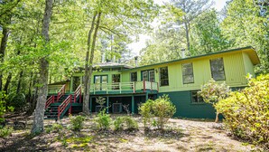 A green wooden cabin with a balcony and a staircase, surrounded by trees.