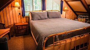 Un dormitorio con techo de madera, una cama con estructura metálica, dos mesitas de noche y un ventanal con cortinas.
