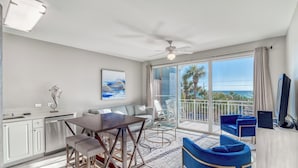 A modern kitchen with white cabinets, a dining area with a wooden table and bar stools, and a balcony with blue chairs and a view of the ocean. - Cabana 30A Gulf Place Cabanas 303 (Santa Rosa Beach)