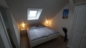 A bedroom with a sloped ceiling, a skylight, a bed with a grey bedspread, two bedside tables with lamps, a door, and a wall-mounted painting. - Neue Ferienwohnung für 2 P. in Sehr Guter Lage! (Borkum)