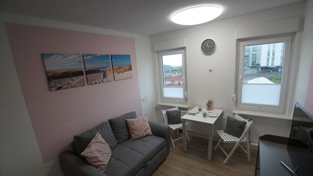 A small, modern room with a grey sofa, a dining table with two chairs, a clock on the wall, and a window with a view of buildings. - Neue Ferienwohnung für 2 P. in Sehr Guter Lage! (Borkum)