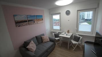 Ein kleiner, moderner Raum mit einem grauen Sofa, einem Esstisch mit zwei Stühlen, einer Wanduhr und einem Fenster mit Blick auf Gebäude.