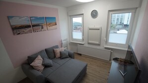 A modern living room with a grey sofa, a flat-screen TV, and a window with a view of buildings. - Neue Ferienwohnung für 2 P. in Sehr Guter Lage! (Borkum)