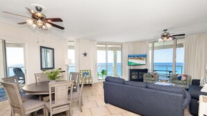 Uma sala de estar espaçosa com um sofá cinza grande, uma mesa de jantar com cadeiras e uma televisão. Há ventiladores de teto, uma decoração de estrela e vista para o oceano através de janelas amplas.
