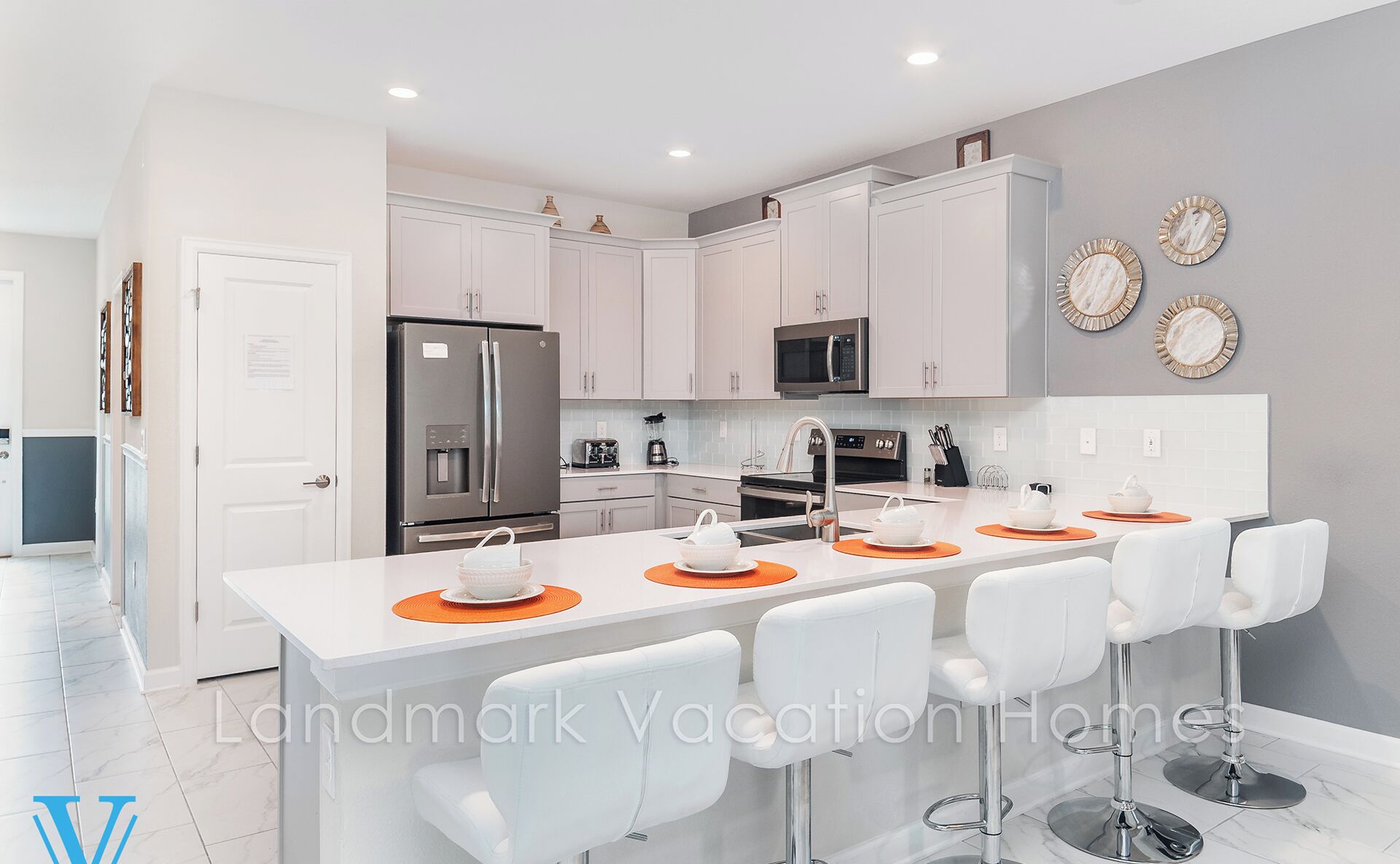 A modern kitchen with white cabinetry, a stainless steel refrigerator, a microwave, and a built-in oven. There are four white bar stools with orange placemats around a central island.