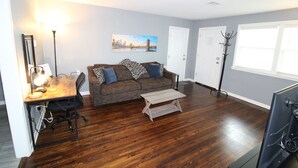 A living room with a wooden floor, a brown sofa with patterned cushions, a small wooden coffee table, a desk with a lamp, and a cityscape painting on the wall. - Recently remodeled house near Expo, TU,  Rt 66, Downtown with large fenced yard (Tulsa)
