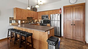 A kitchen with wooden cabinets, a stainless steel refrigerator, a microwave, and a sink. There are three bar stools and a trash can.