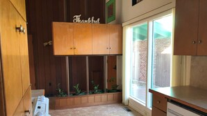A kitchen with wooden cabinets, a stone-tiled floor, and a window with a view of a brick wall. - Sunshine House (Ellis)