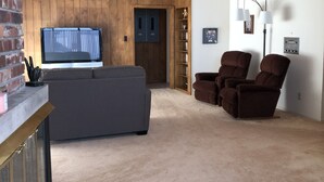 A living room with a large grey sofa, two brown recliners, a TV, and a fireplace. - Sunshine House (Ellis)