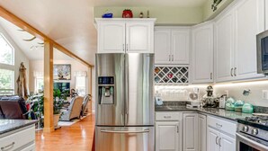 Una cocina moderna con armarios blancos, electrodomésticos de acero inoxidable y un comedor con sofá de cuero y plantas.