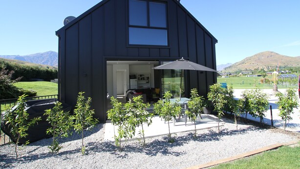 A modern black house with a large window, a covered patio, and a gravel driveway.