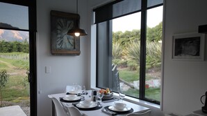 A dining area with a table set for two, a clock on the wall, and a view of a garden through large windows.