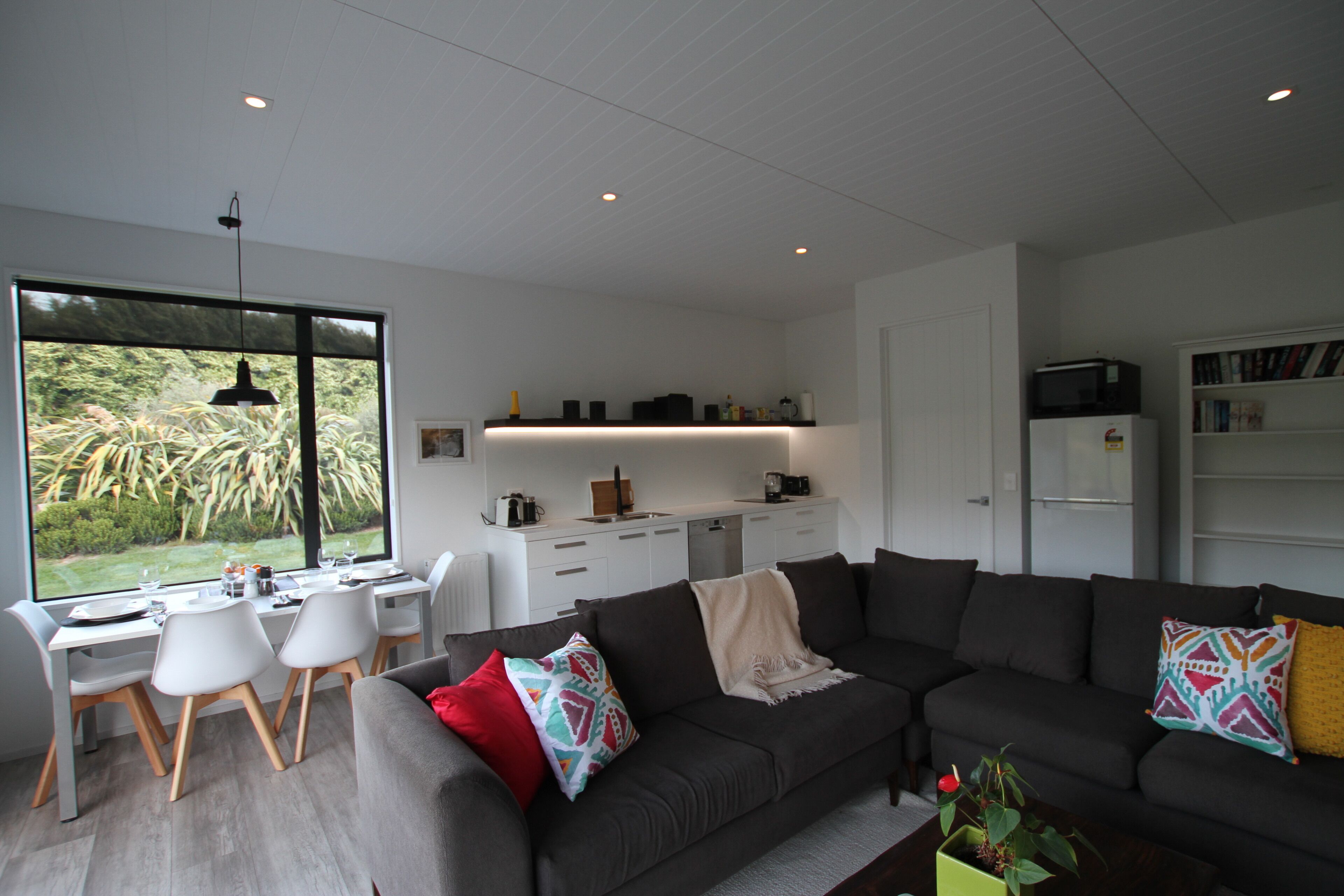 A modern living room with a dark grey sofa, a dining area with white chairs, and a kitchen with white cabinets.