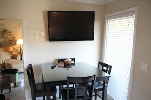 Un comedor con una mesa de vidrio, cuatro sillas, un televisor de pantalla plana montado encima y una ventana con persianas.