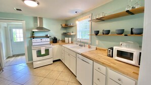 A kitchen with white cabinets, a stove, a dishwasher, and a microwave. There are shelves with bowls and glasses, and a window with blinds.