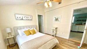 A bedroom with a bed, bedside table, and a nightstand. There is a door leading to a bathroom and a kitchen.