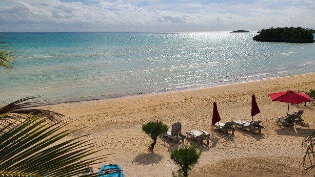 Una playa con agua turquesa cristalina, una orilla arenosa y una pequeña isla a lo lejos. Hay sillones de playa y sombrillas dispuestas para el descanso.