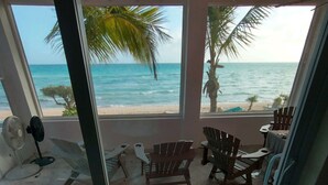 Habitación con vistas a la playa y al océano a través de amplios ventanales. Varias sillas de madera y una pequeña mesa.