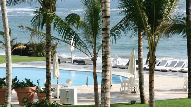 Un espace piscine agrémenté de chaises longues et de parasols, entouré de palmiers et offrant une vue sur l’océan.