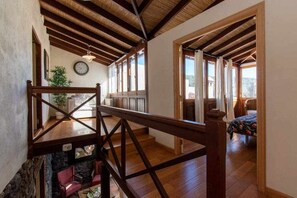 Um quarto espaçoso com vigas de madeira no teto, uma cama com colcha florida e vista para o exterior através de grandes janelas.