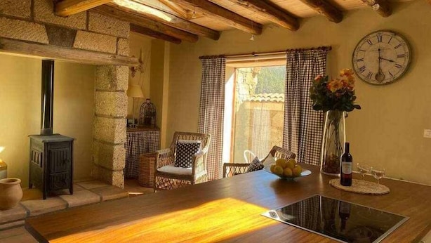 A rustic kitchen with exposed wooden beams, a stone wall, a wooden stove, a dining table with chairs, and a clock on the wall.