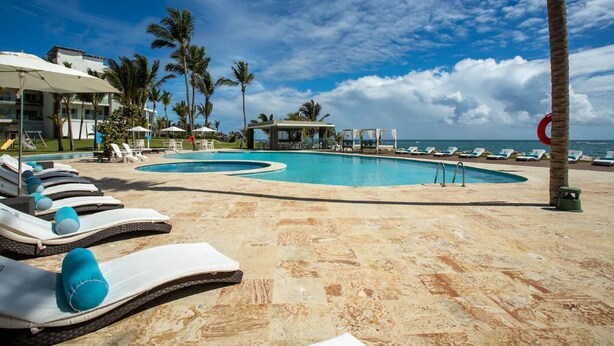 Un espace piscine doté de chaises longues et de parasols, de palmiers, et offrant une vue sur l’océan.