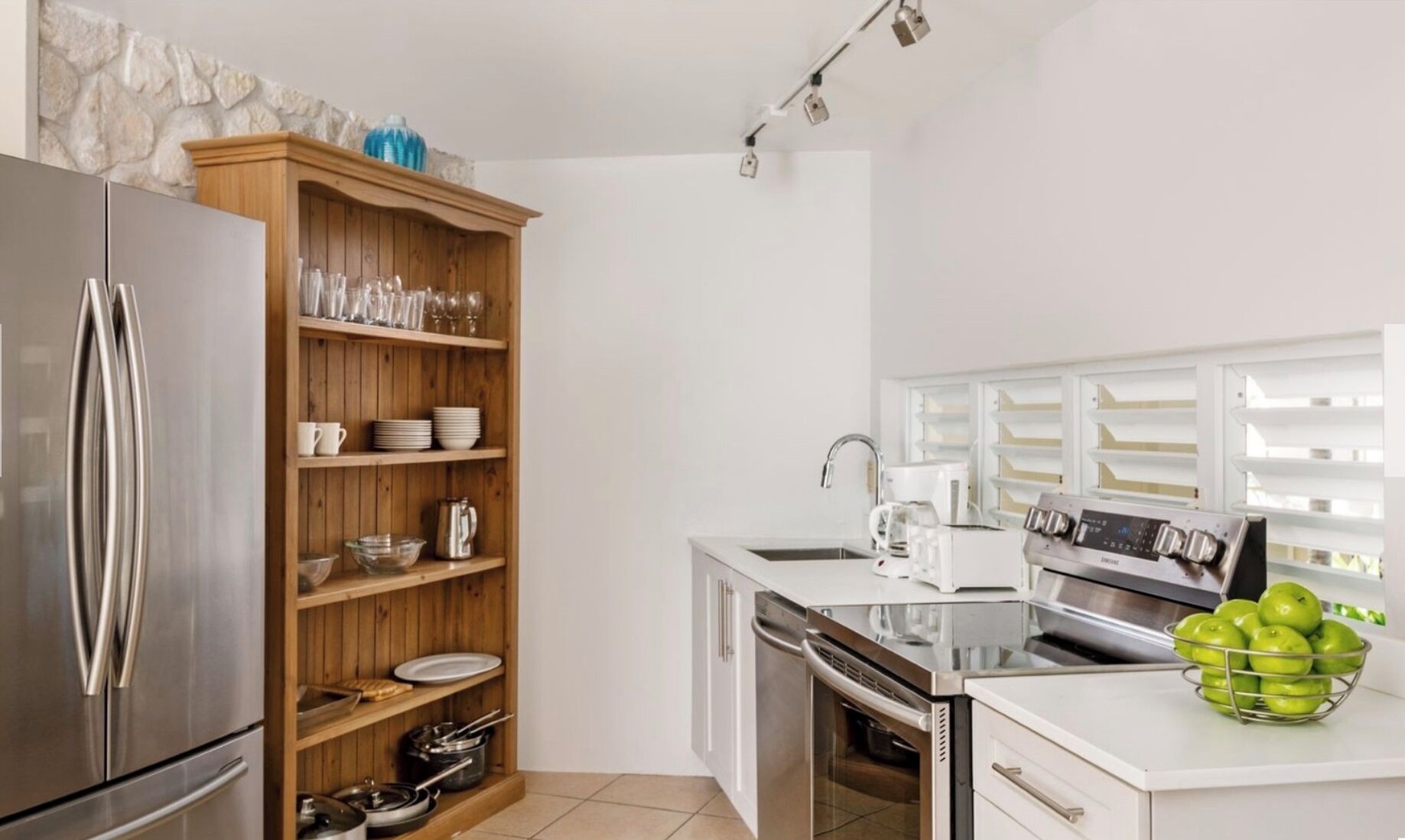 Una cocina moderna con un refrigerador de acero inoxidable, repisas de madera con vajilla, un horno empotrado y una encimera blanca con una cafetera y un frutero.