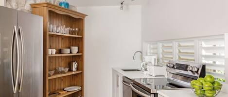 Una cocina moderna con un refrigerador de acero inoxidable, repisas de madera con vajilla, un horno empotrado y una encimera blanca con una cafetera y un frutero.