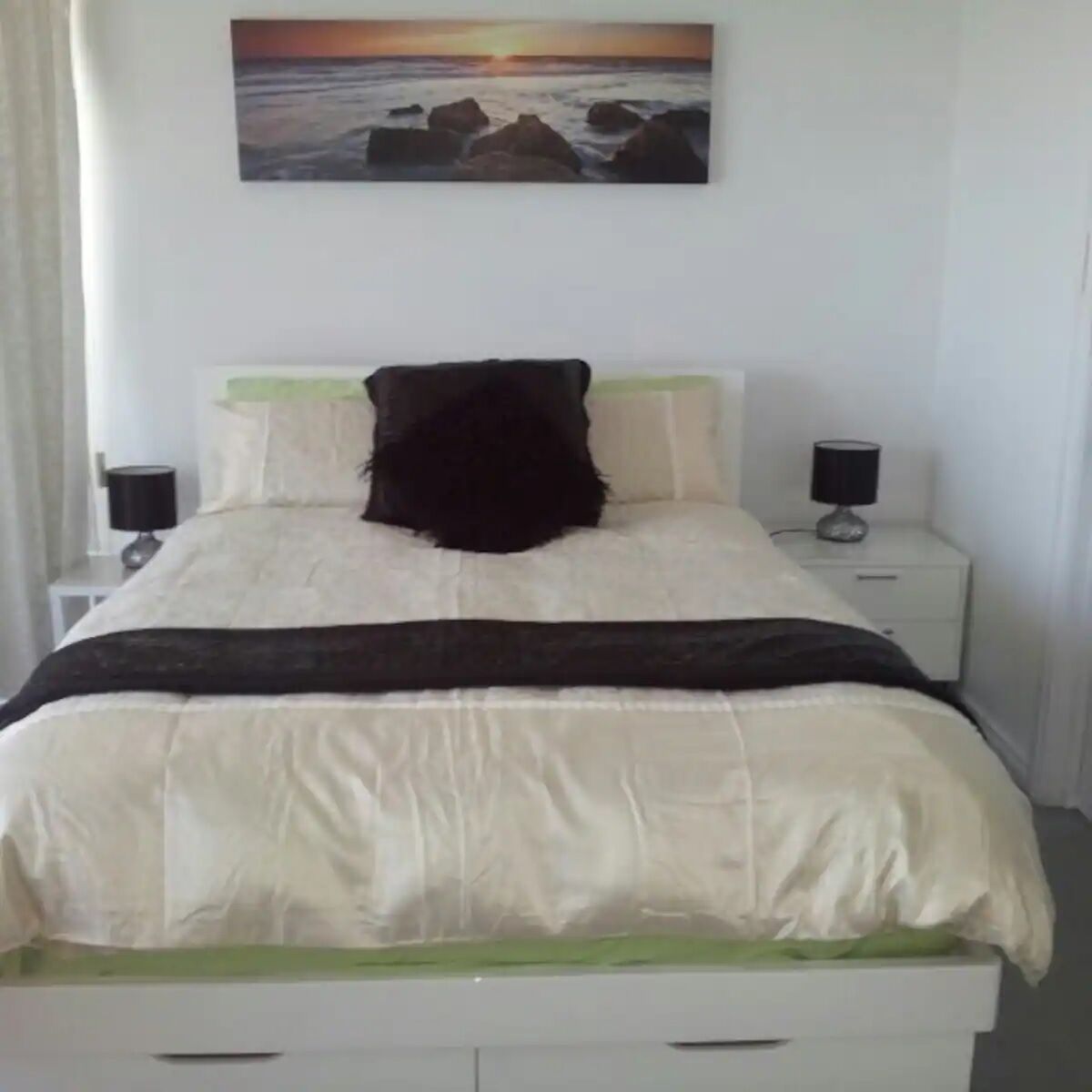 A neatly made bed with a white and black bedspread, two bedside lamps, and a painting of a sunset over rocks on the wall.