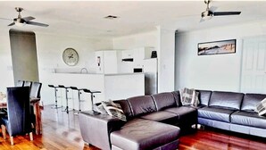 Una sala moderna con un sofá modular, un comedor con sillas, una cocina al fondo y un ventilador de techo.