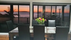 Un comedor con mesa y sillas, ventanas amplias con vista al atardecer sobre el agua, y una planta en maceta sobre la mesa.