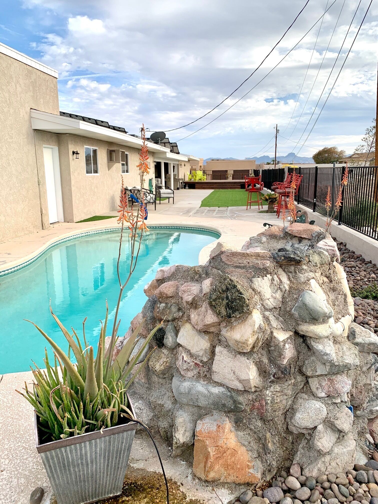 Una piscina con pared de piedra y plantas, un patio con mobiliario exterior y vistas a una cadena montañosa.