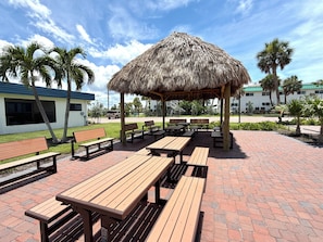 An outdoor seating area with wooden benches and picnic tables, a thatched-roof pavilion, and palm trees.
