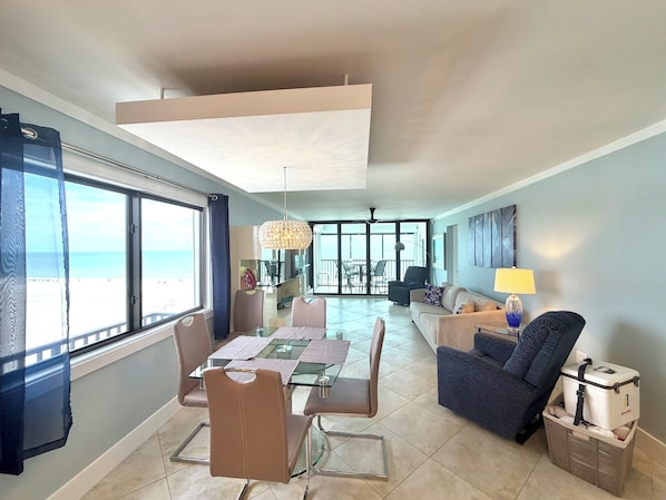 A spacious living area with a dining table, chairs, a sofa, and a view of the sea through large windows.