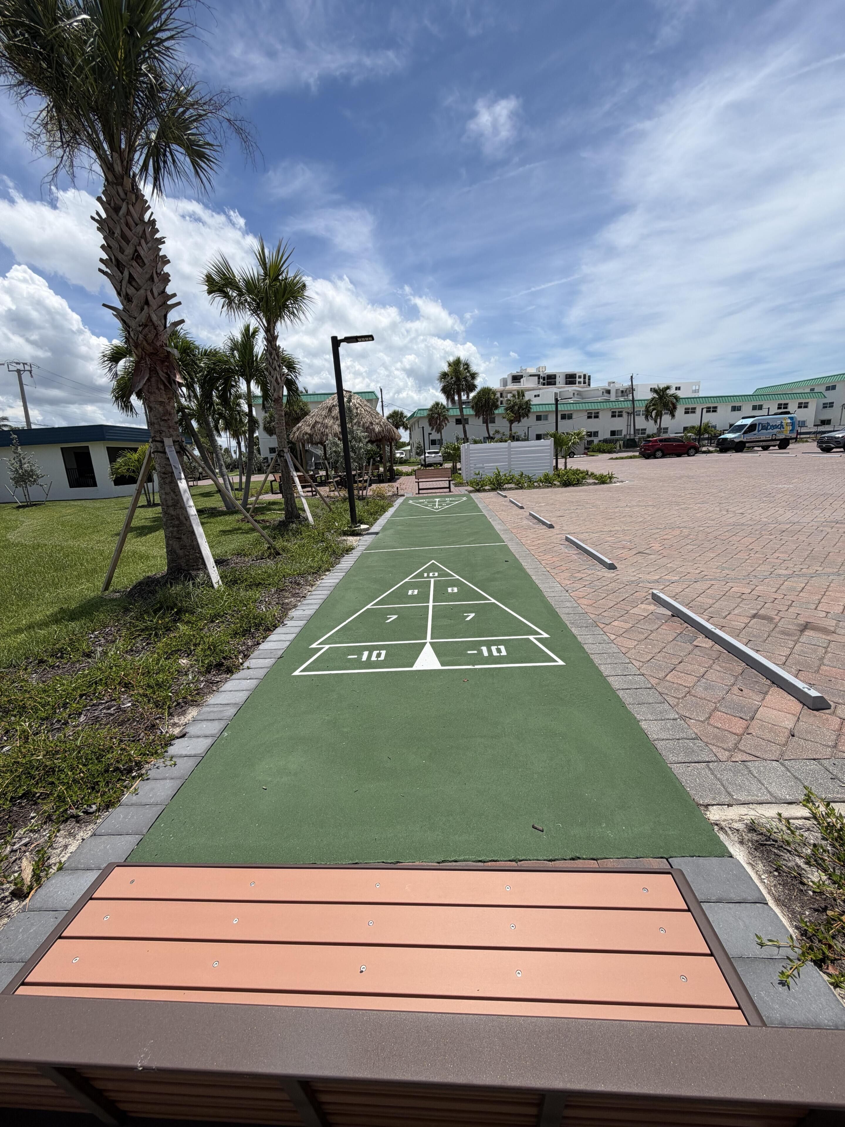 Un sendero verde con un juego de rayuela, palmeras y un estacionamiento con un edificio al fondo.