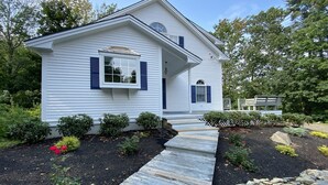 Une maison de plain-pied avec un porche à l’avant, un petit jardin avec différentes plantes et fleurs, et un mur en pierre.