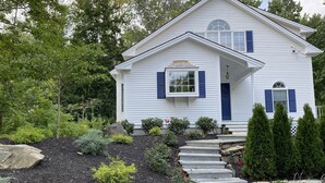 Une maison blanche aux volets bleus, un escalier en pierre et un jardin où poussent différentes espèces de plantes.