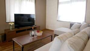 A living room with a flat-screen TV on a wooden stand, a glass coffee table with wine glasses and a fruit basket, a white sofa, and large windows with sheer curtains. - Pool Villa Sesoko jima (Motobu)