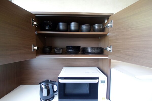 A compact kitchen with a microwave and kettle, shelves with black bowls and plates, and wooden cabinetry. - Pool Villa Sesoko jima (Motobu)