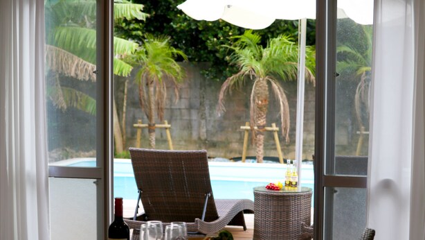 A room with a glass table, a wicker chair, a wicker side table, a bottle of wine, and a fruit basket. - Pool Villa Sesoko jima (Motobu)
